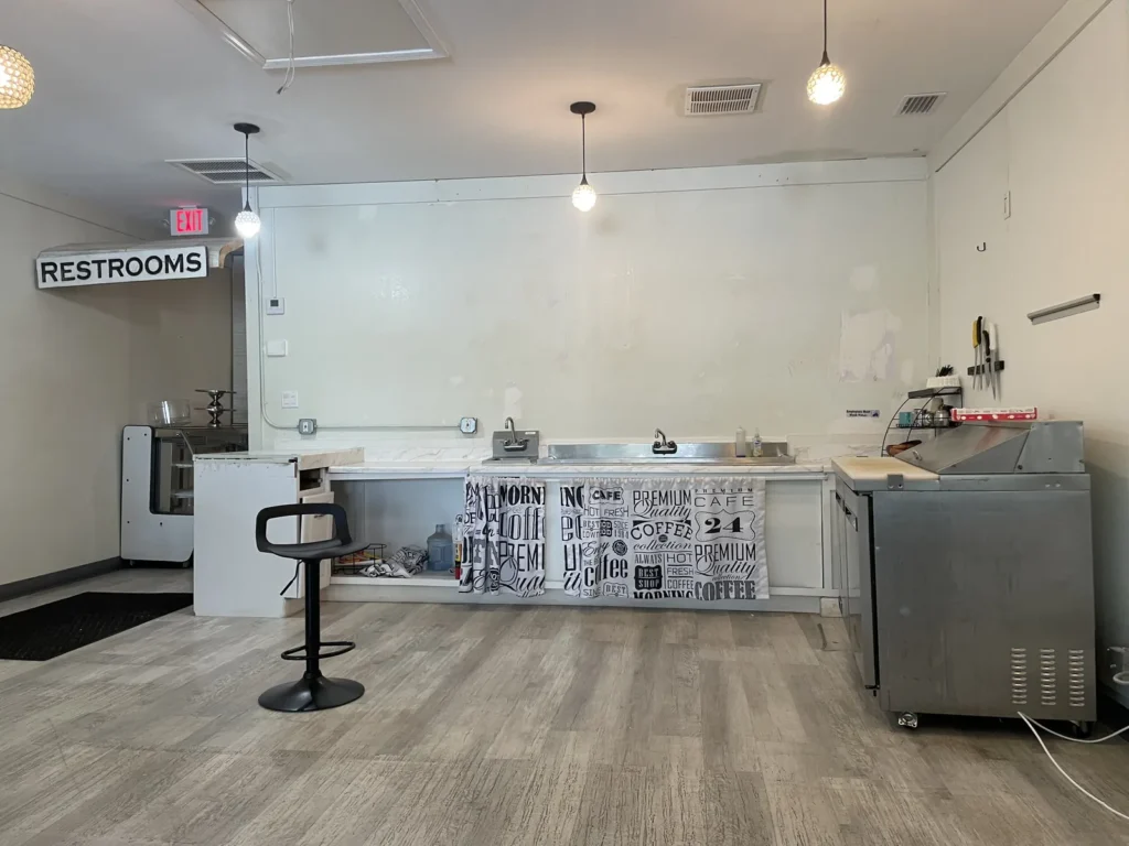 Espacio de cafetería antes de la remodelación, con pared blanca parchada, mostrador con fregadero y letrero de baños