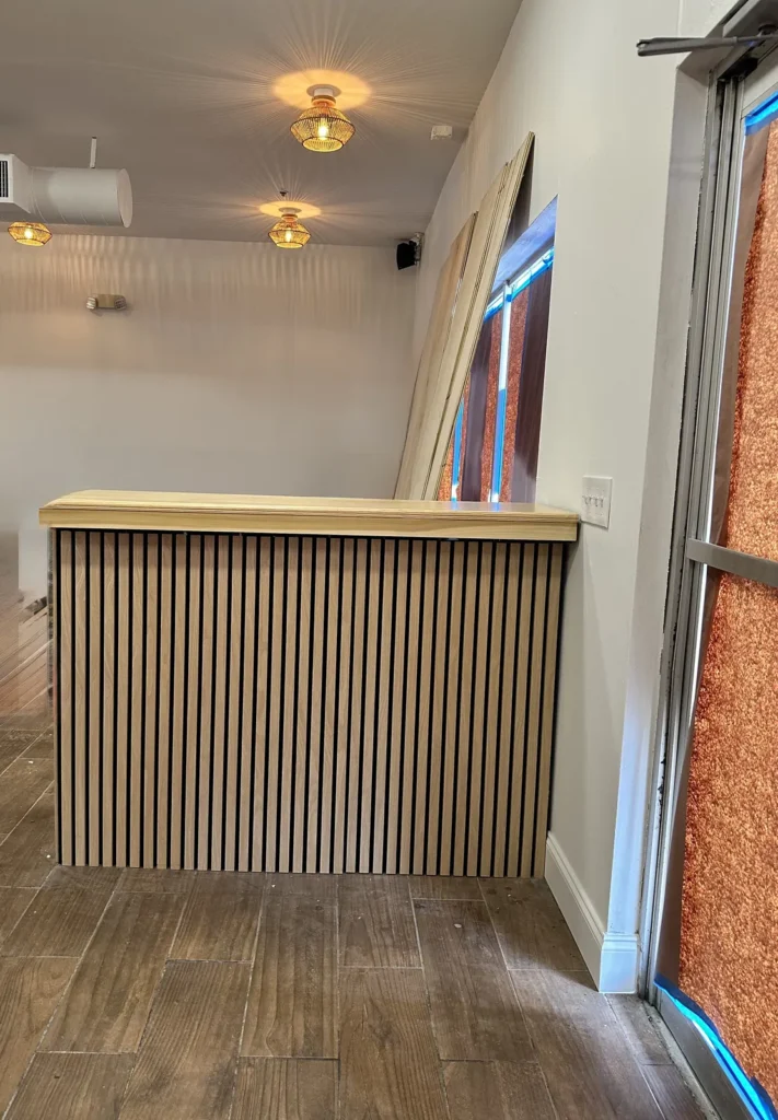Finished commercial reception counter with a natural wood slat front, wood top, rattan pendant lights, and wood-look tile floor